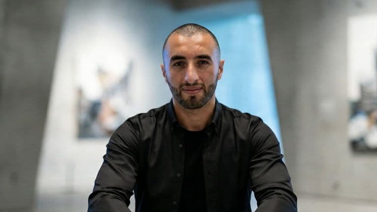 Man in a black shirt with a confident expression stands in a modern art gallery, surrounded by abstract paintings on concrete walls.