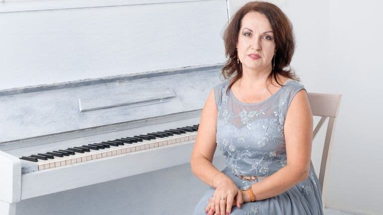 A woman in a blue dress sits on a chair beside a white upright piano, conveying a calm and composed atmosphere. The room is bright and minimalistic.