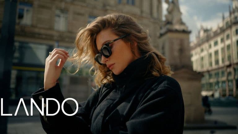 A woman in sunglasses and a black coat stands in a city square, with elegant architecture behind her. She looks contemplative. The word "Lando" is displayed.