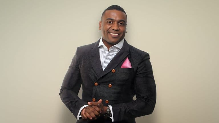Man in a textured dark suit with a pink pocket square, smiling warmly against a light beige background. His hands are clasped in front.