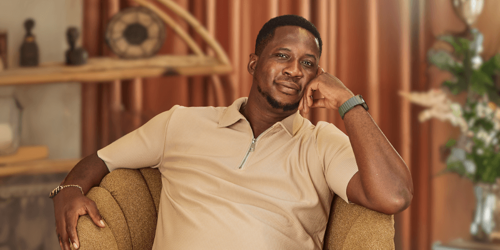 "A confident man in a beige polo shirt smiles while resting his head on his hand, seated comfortably on a stylish armchair in a modern, cozy interior with soft lighting and decorative plants."