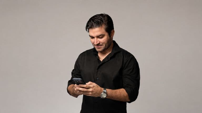 Man in black shirt looking at smartphone, wearing a wristwatch, against a neutral studio background.