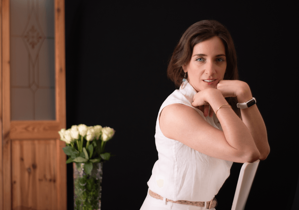Portrait of a woman dressed in a white sleeveless top, posing confidently with a bouquet of white roses in the background.