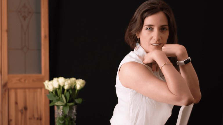 Portrait of a woman dressed in a white sleeveless top, posing confidently with a bouquet of white roses in the background.