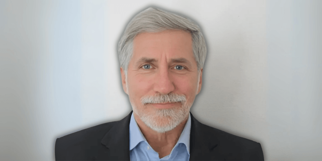 Portrait of a man with gray hair and a beard, dressed in a blue shirt and black blazer, smiling in front of a neutral background.