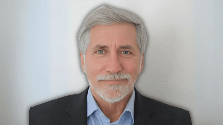 Portrait of a man with gray hair and a beard, dressed in a blue shirt and black blazer, smiling in front of a neutral background.