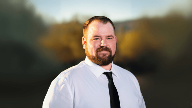 Portrait of a man with a full beard, dressed in a white shirt and black tie, standing outdoors with a blurred natural background. He looks directly at the camera with a neutral expression.