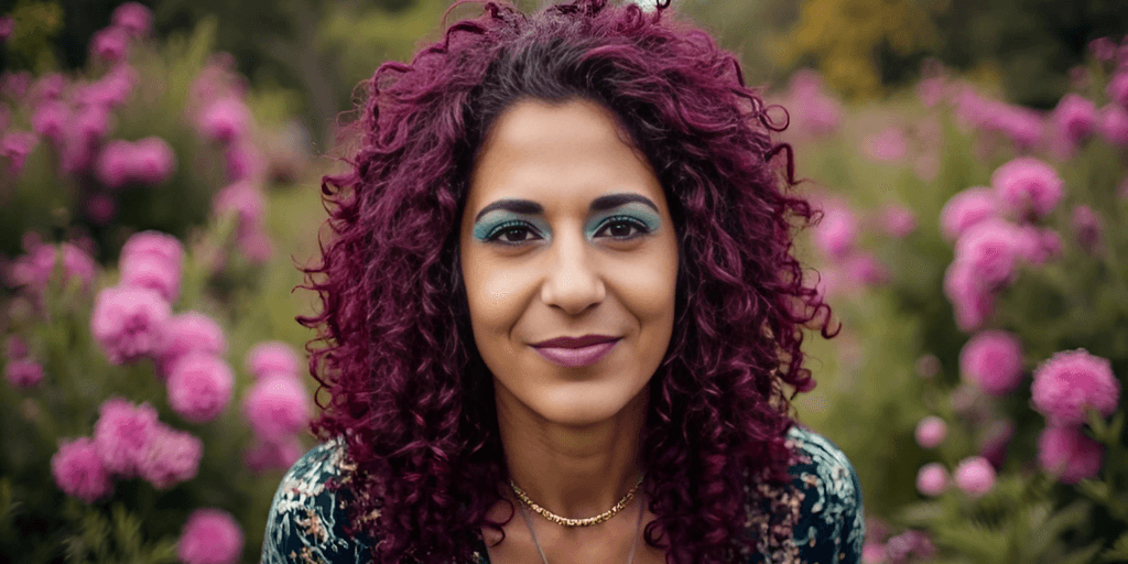 A woman with curly purple hair and blue eyeshadow, smiling in front of a background of pink flowers.