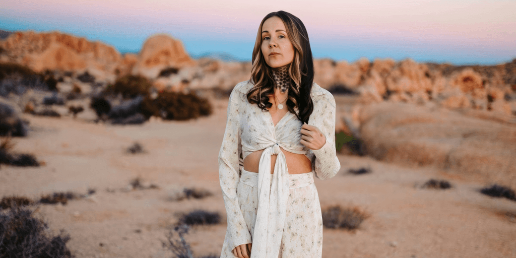 A woman stands in a desert landscape, wearing a white flowy outfit, with a calm expression