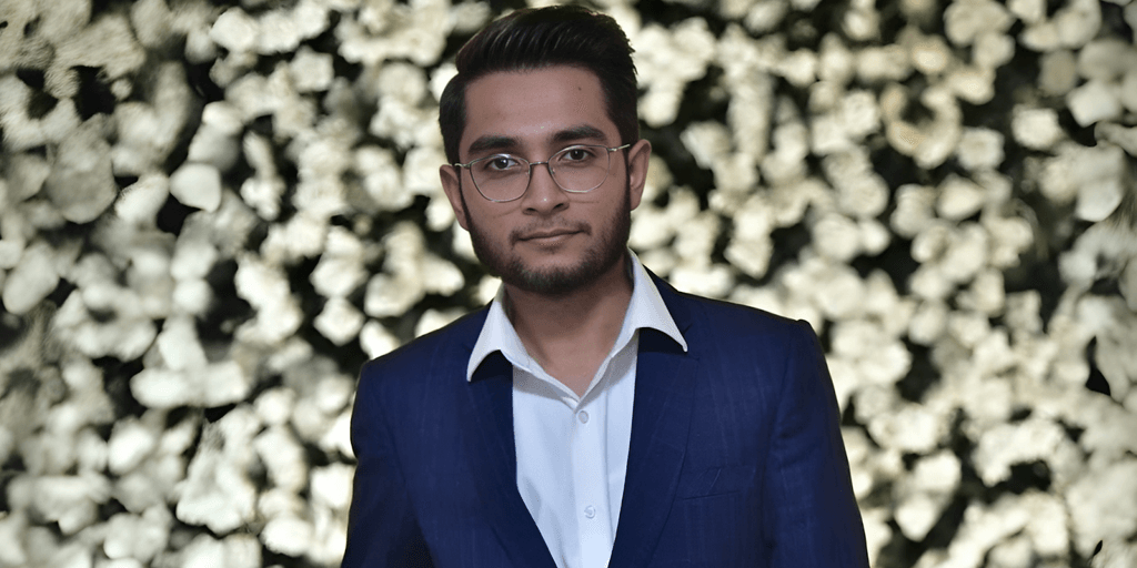Man in a blue suit with glasses, standing in front of a flower wall.