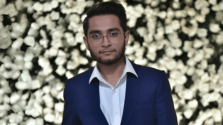 Man in a blue suit with glasses, standing in front of a flower wall.