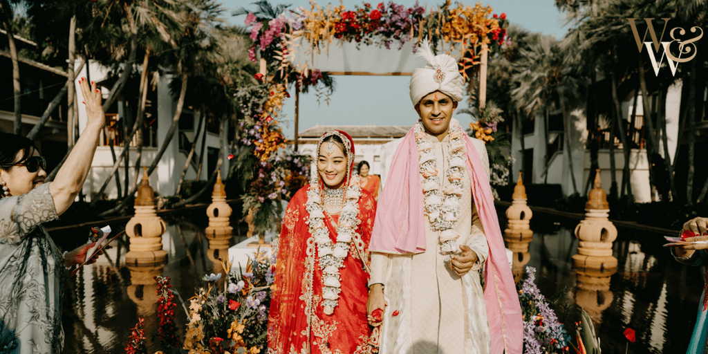 Indian couple dressed in traditional wedding attire, walking together at their ceremony surrounded by a lush floral arch, with family members celebrating in the background.