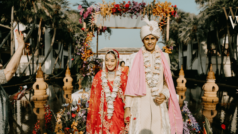 Indian couple dressed in traditional wedding attire, walking together at their ceremony surrounded by a lush floral arch, with family members celebrating in the background.