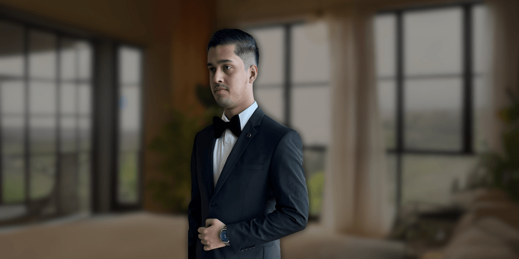 Man in formal black tuxedo and bow tie standing in a well-lit modern living room with large windows, showcasing a sophisticated look.