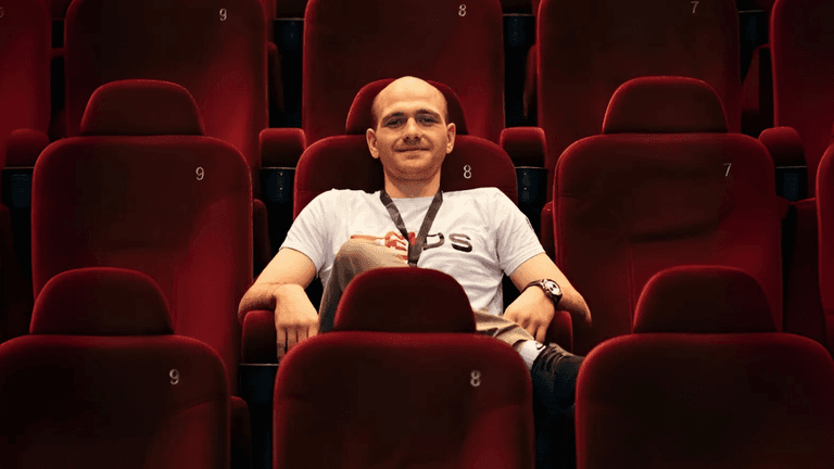 Person relaxing in a cinema chair, wearing a M3DS Academy t-shirt, in an empty movie theater with vibrant red seats.