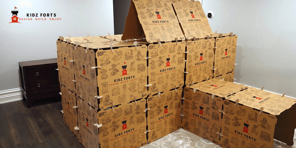 A large, creative fort built from Kidz Forts cardboard building kits, featuring multiple stacked boxes with playful designs and illustrations. The fort has a pitched roof and is designed for children to build and enjoy.