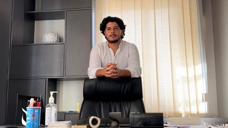 A man standing in a modern office space, wearing a white shirt and smiling, with his hands resting on the desk, looking confident.
