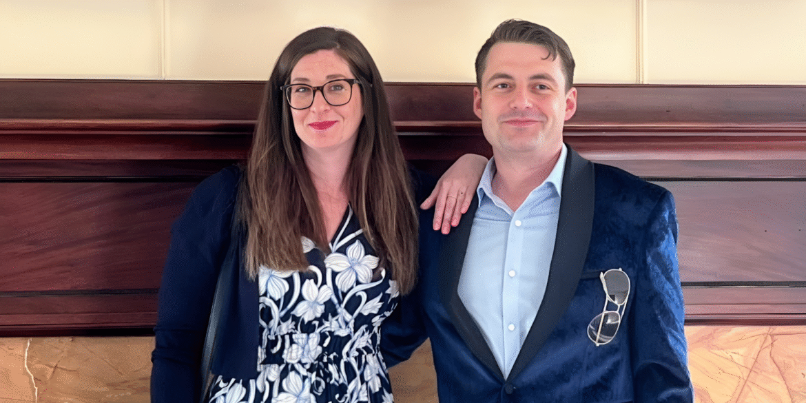 Man and woman smiling in front of wooden panel, dressed in business attire.