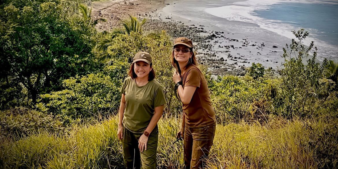 Two women hiking through lush greenery with a scenic beach view in the background.