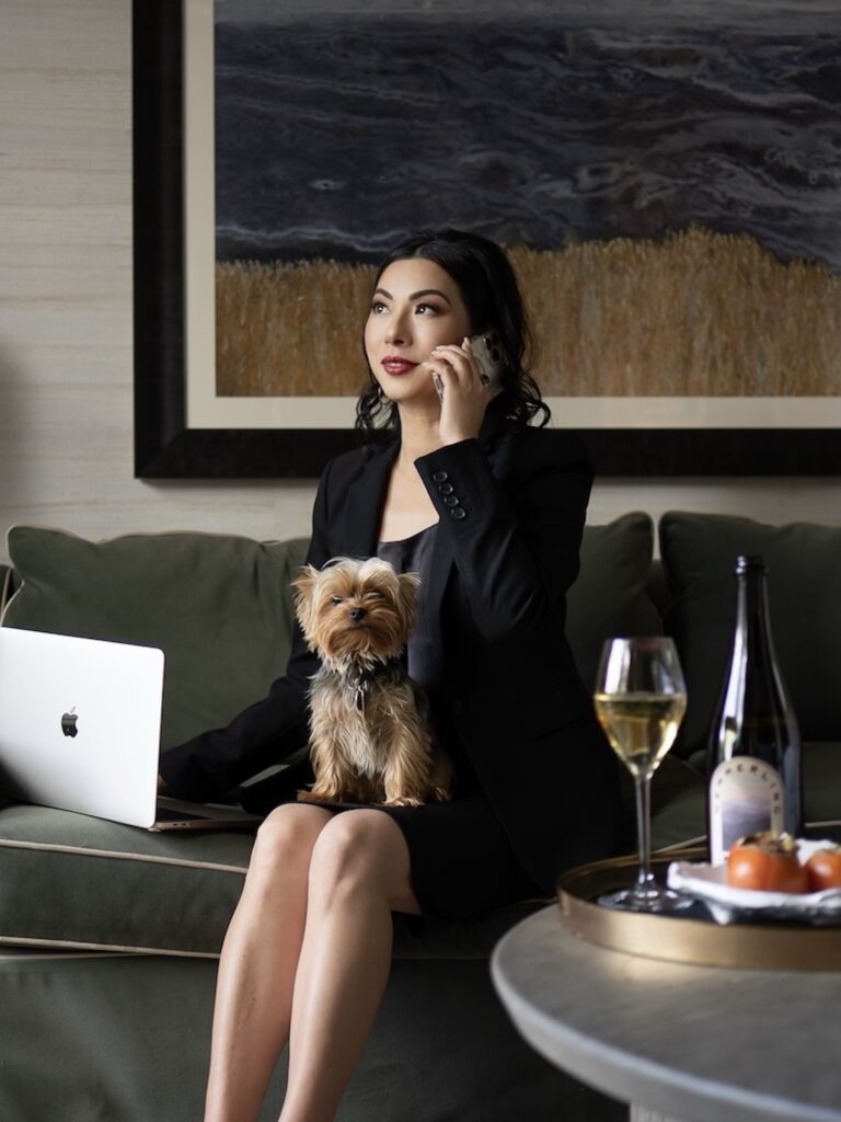 Susye Weng-Reeder working remotely from a hotel suite with her Yorkie Einstein on her lap, glass of wine nearby—balancing thought leadership with luxury lifestyle.