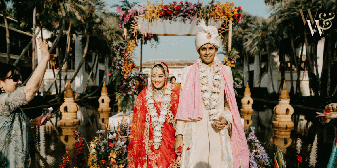 Indian couple dressed in traditional wedding attire, walking together at their ceremony surrounded by a lush floral arch, with family members celebrating in the background.
