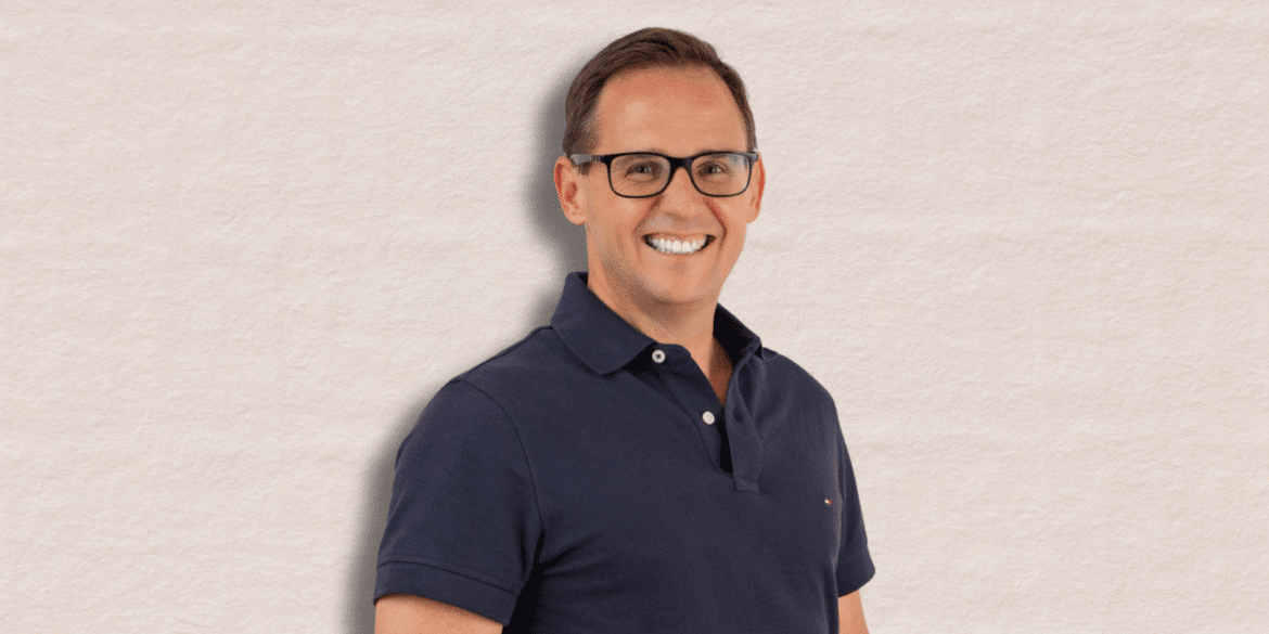 Smiling professional man in a navy polo shirt with glasses, standing against a soft beige background, exuding confidence and approachability.