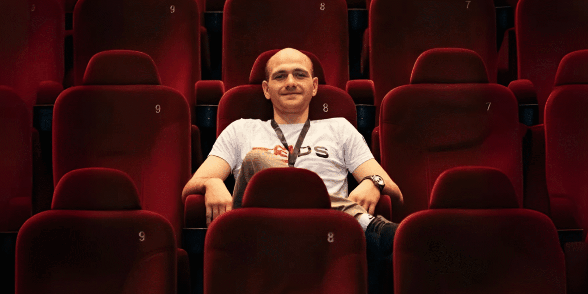 Person relaxing in a cinema chair, wearing a M3DS Academy t-shirt, in an empty movie theater with vibrant red seats.