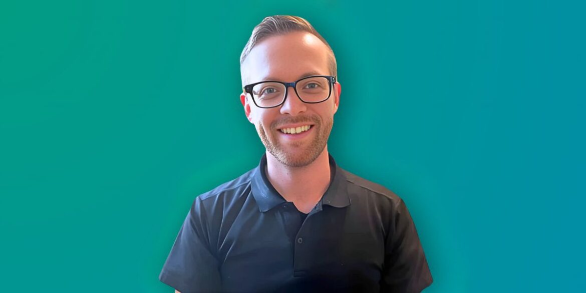 Portrait of a smiling man wearing glasses and a black polo shirt, standing against a vibrant teal background.