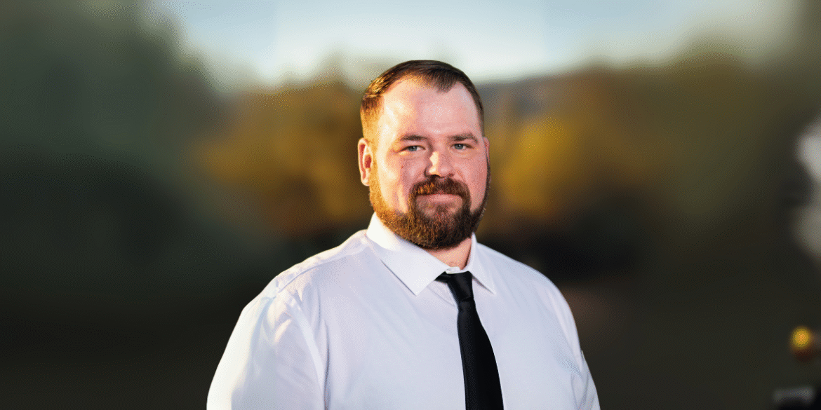 Portrait of a man with a full beard, dressed in a white shirt and black tie, standing outdoors with a blurred natural background. He looks directly at the camera with a neutral expression.