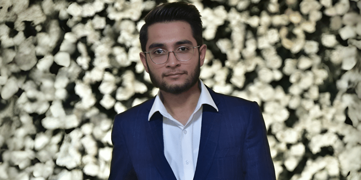 Man in a blue suit with glasses, standing in front of a flower wall.
