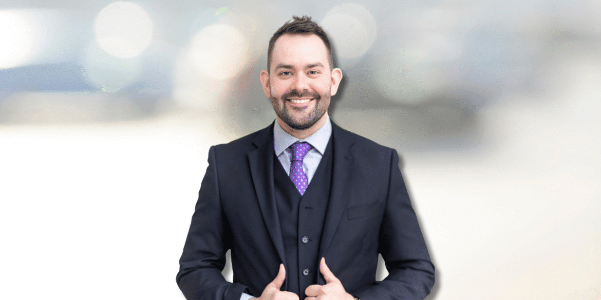 Professional portrait of a man in a suit with a purple tie and a confident smile. He stands with his thumbs up, set against a blurred background of soft lights, portraying a confident and approachable business image.