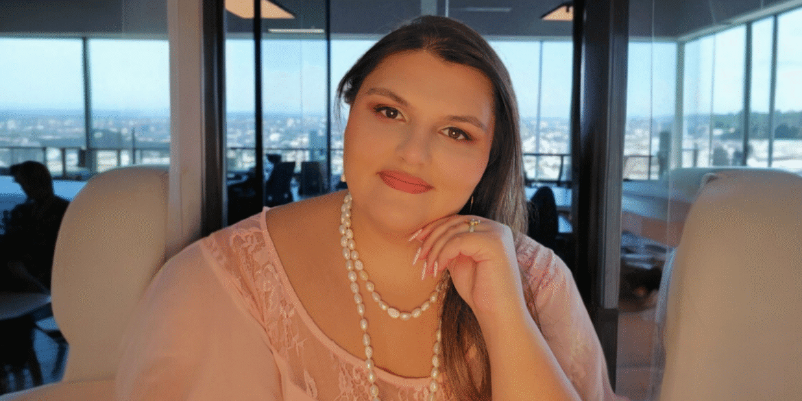 A woman with long hair and pearl jewelry, sitting and smiling while posing with her hand on her chin. She is in a modern office with a panoramic view of the city in the background.