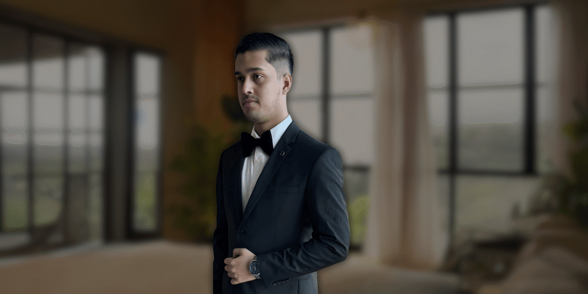 Man in formal black tuxedo and bow tie standing in a well-lit modern living room with large windows, showcasing a sophisticated look.