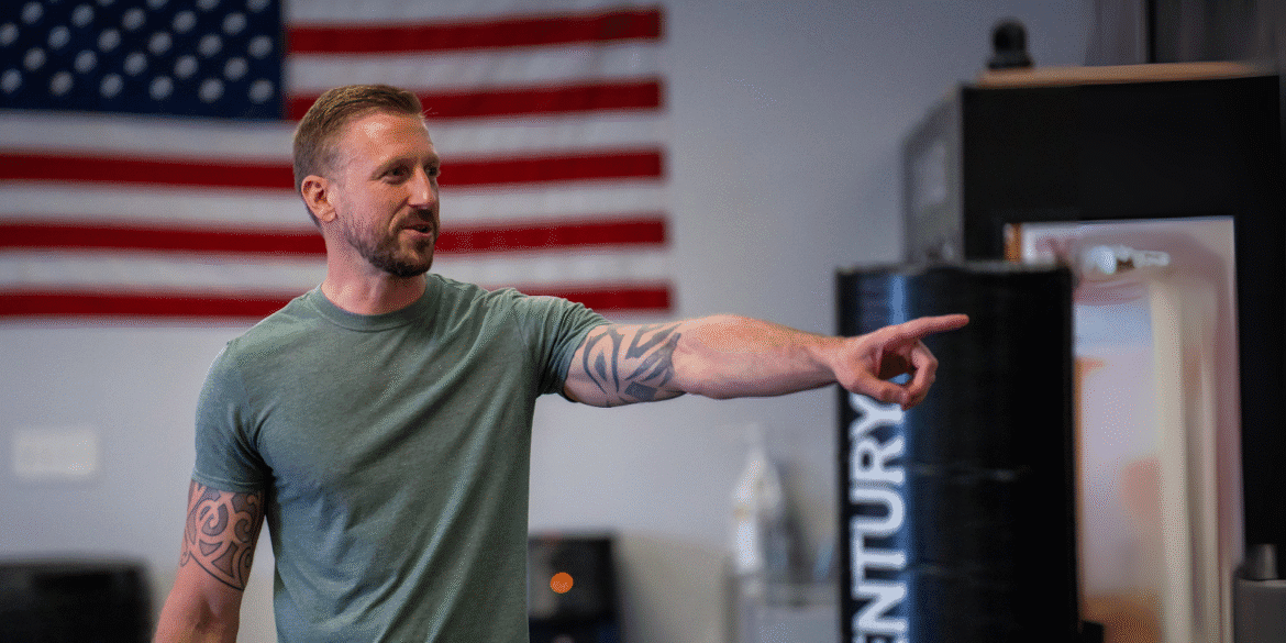 Man with tattoos pointing while speaking, with American flag in background.