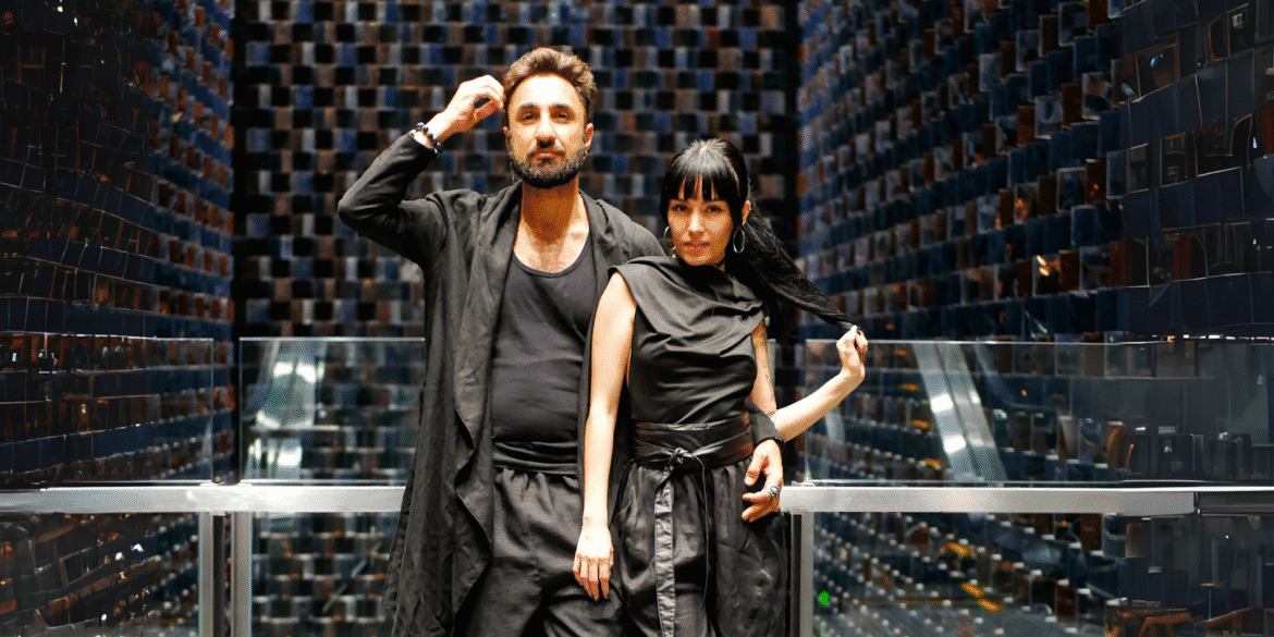 A stylish couple posing in black outfits against a backdrop of mirrored walls with a modern, geometric design.