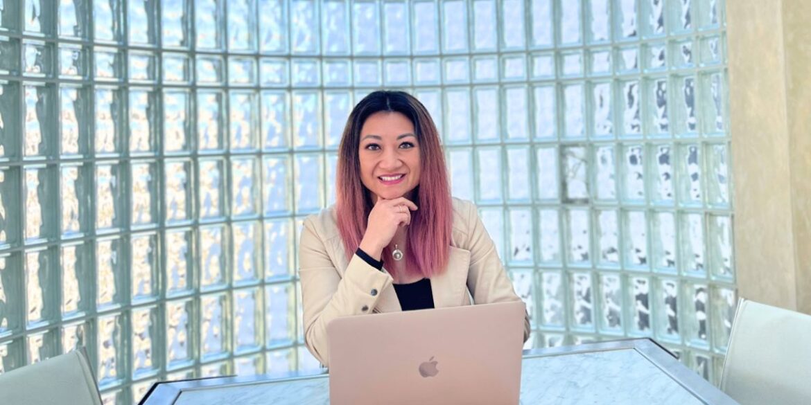 "Professional portrait of a woman with pink ombre hair, sitting at a desk with a MacBook, smiling and resting her chin on her hand, against a backdrop of a glass block wall."