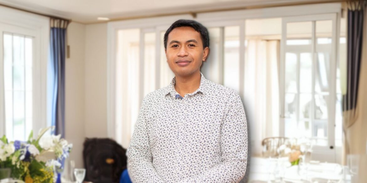 Man in a floral shirt standing indoors with a soft smile, surrounded by natural light and a blurred background of curtains and floral arrangements.