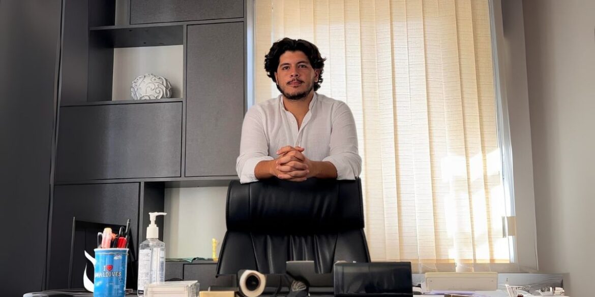 A man standing in a modern office space, wearing a white shirt and smiling, with his hands resting on the desk, looking confident.
