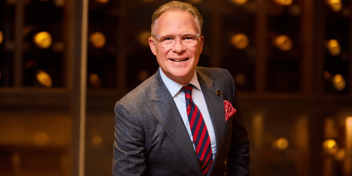 A confident man in a tailored suit stands smiling in front of a wine cellar, showcasing presence and leadership.