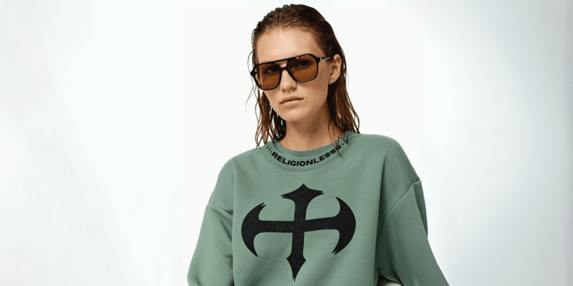 "Model wearing a green sweatshirt with the Religionless logo on the front and bold sunglasses, posing confidently against a simple white background."