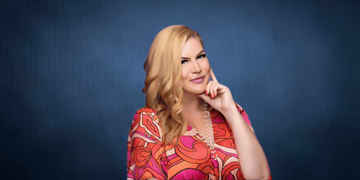 Professional portrait of a woman in a colorful patterned blouse, smiling and resting her chin on her hand with a confident expression against a blue background.