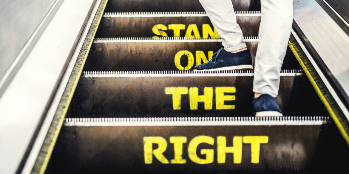 Person walking up an escalator with the words 'STAND ON THE RIGHT' clearly visible in yellow on the steps.