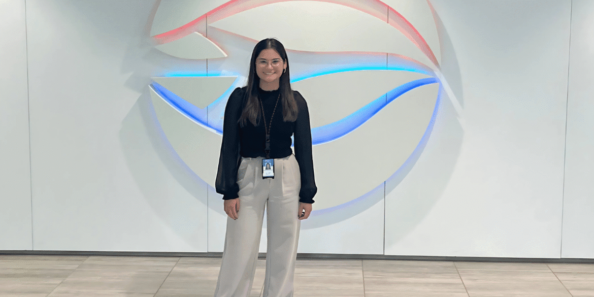 Professional young woman smiling confidently in front of a modern, illuminated company logo, dressed in business attire with light-colored trousers and black top."