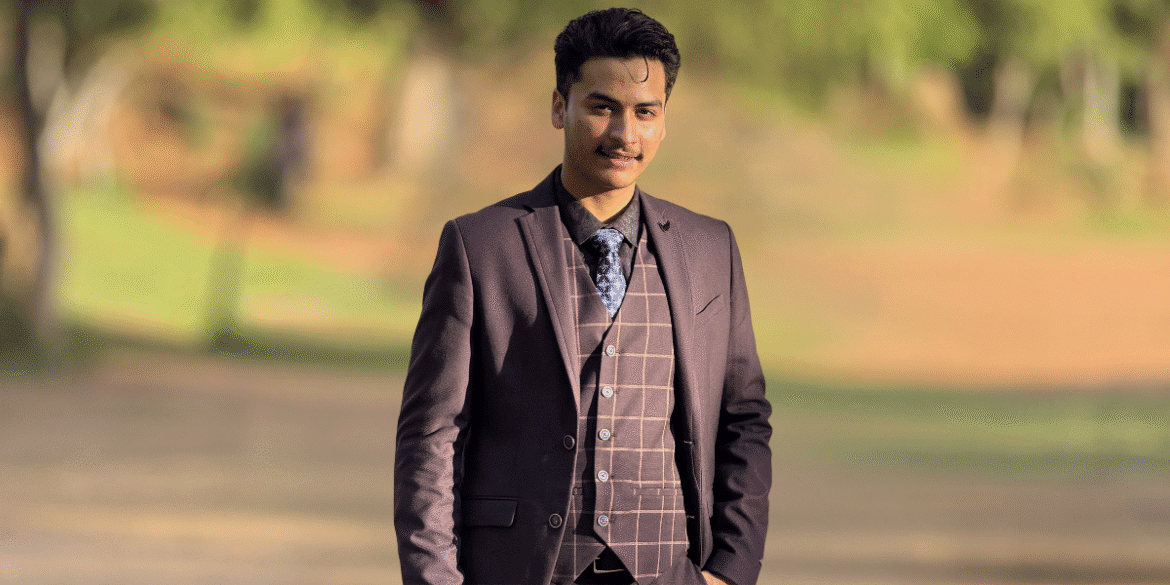 "Young man in a brown suit with a plaid pattern, standing confidently in a park with blurred greenery in the background, showcasing a professional appearance."