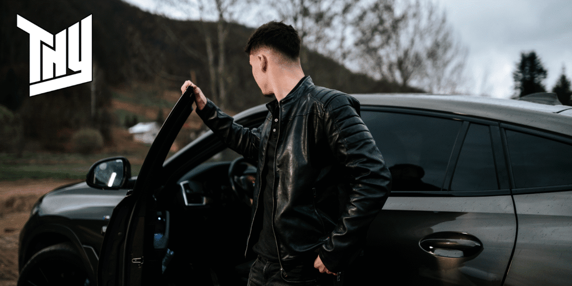 "Young man in a black leather jacket standing next to a car, opening the door while looking away, with a scenic background of hills and trees."