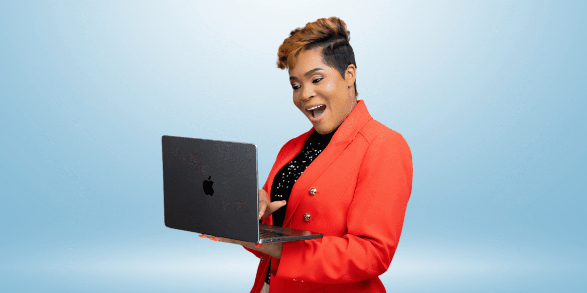 Woman in a red blazer happily using a laptop with a bright blue background, displaying enthusiasm and engagement.