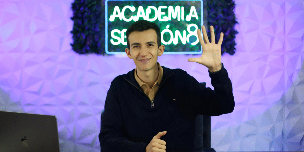 Smiling young man wearing a navy sweater and beige shirt, sitting at a desk with a MacBook in front of him. He is raising his right hand with five fingers up, posing in front of a neon sign that reads 'Academia Secreta' with a modern geometric background.