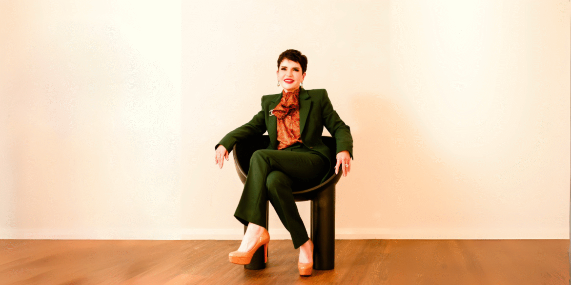 Portrait of a confident woman in a green suit, sitting in a modern chair with high heels, looking relaxed.