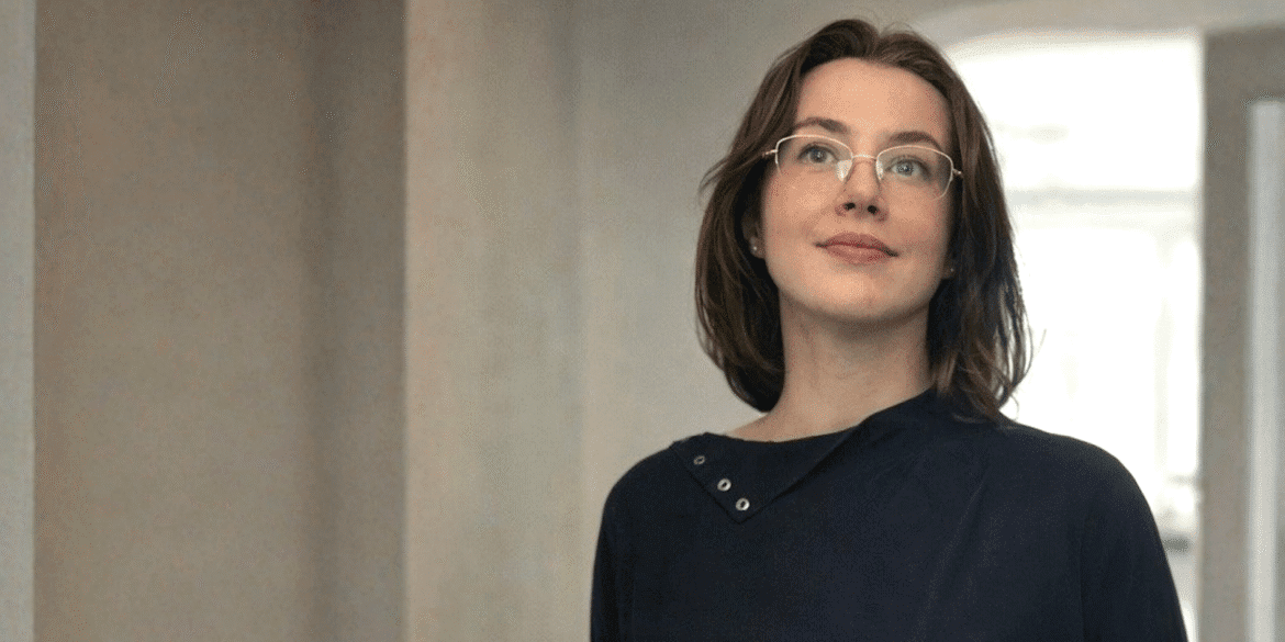 Blonde woman with blue eyes wearing a black blouse and leather vest against a textured beige wall.