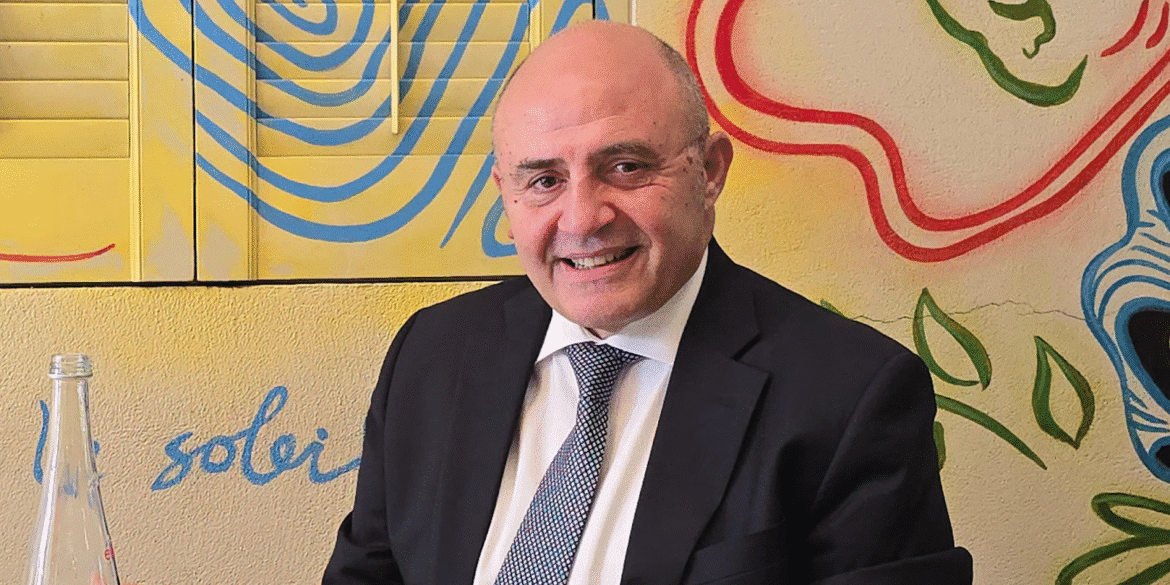 "Man seated at a table with a colorful mural in the background, wearing a dark suit and a patterned tie. He smiles warmly, exuding a relaxed and approachable demeanor."